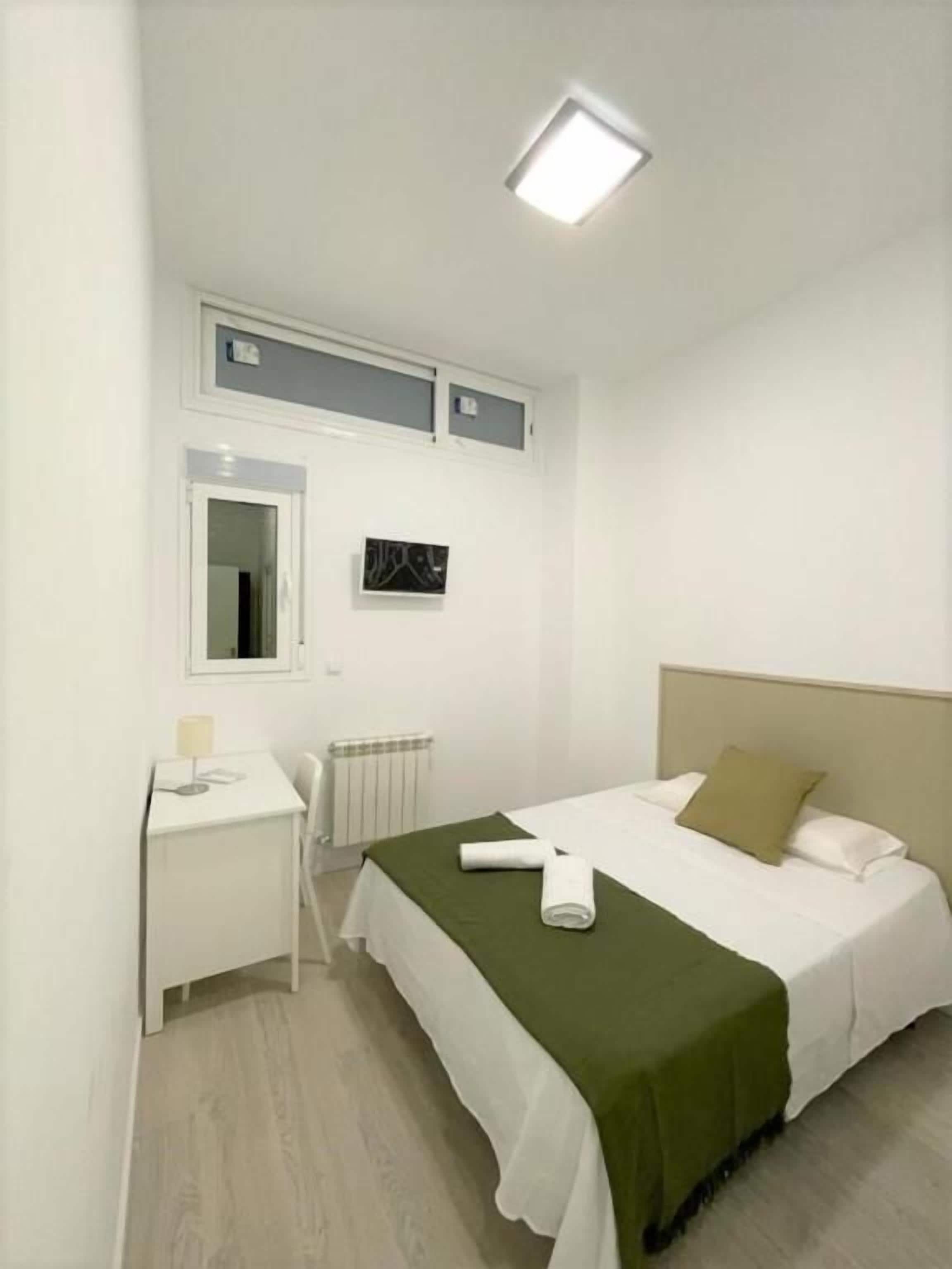 A hotel room with a bed, a desk, a chair, a radiator, and a ceiling light.