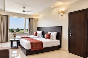 Habitación de hotel con una cama grande, una mesita de noche y una puerta. Tiene cortinas en la ventana y un ventilador de techo.