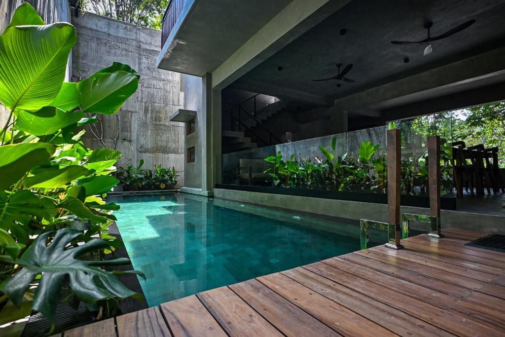 A modern outdoor pool area with a wooden deck, surrounded by lush greenery and a concrete structure.