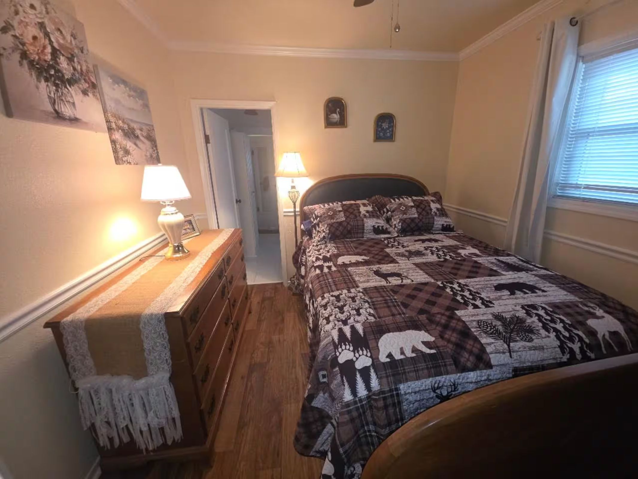 A bedroom with a bed, dresser, two lamps, and a door leading to another room.