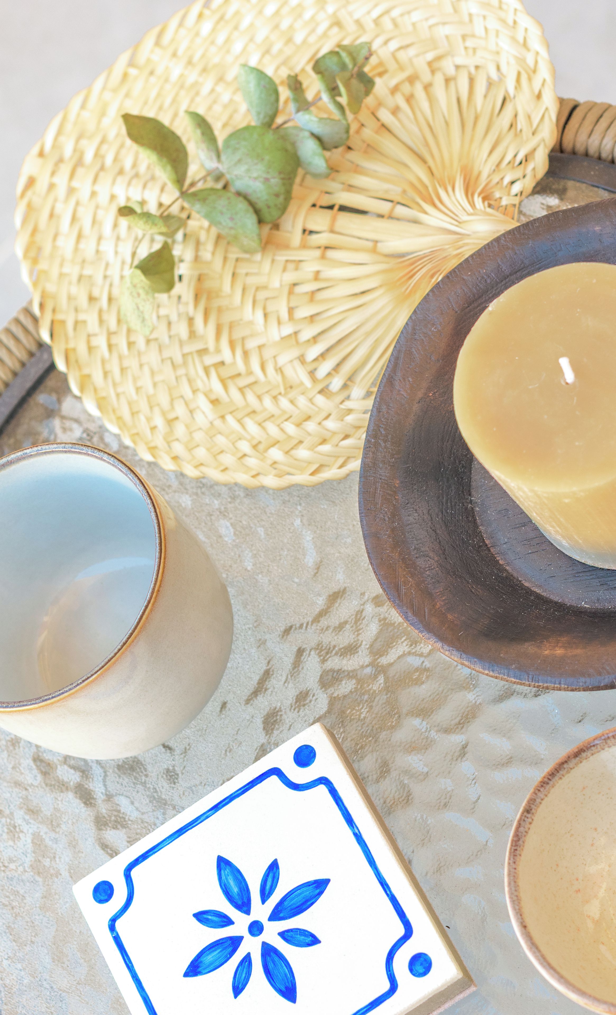 A tray with a woven basket, a candle, a ceramic cup, and a decorative tile.