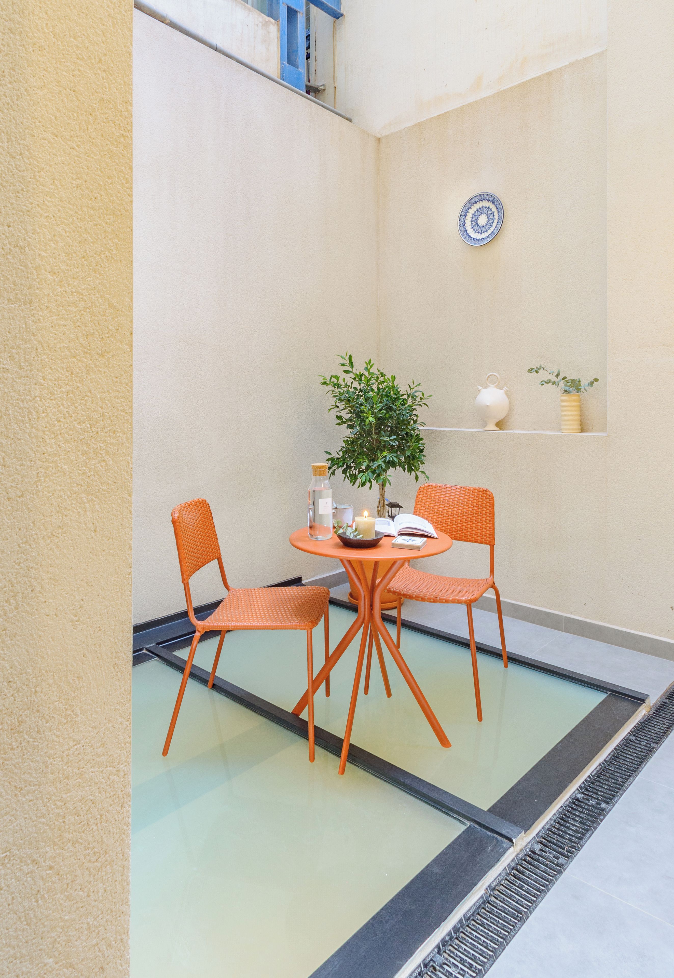 Un petit espace extérieur aménagé avec deux chaises orange, une table ronde, une plante en pot et une assiette décorative accrochée au mur.