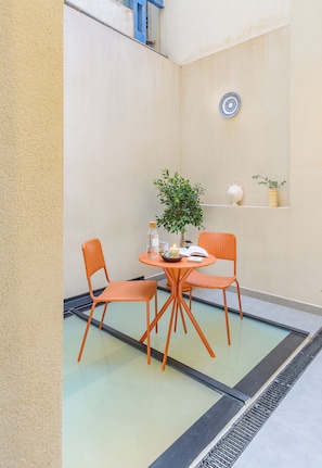 A small outdoor seating area with two orange chairs, a round table, a potted plant, and a decorative plate on the wall.