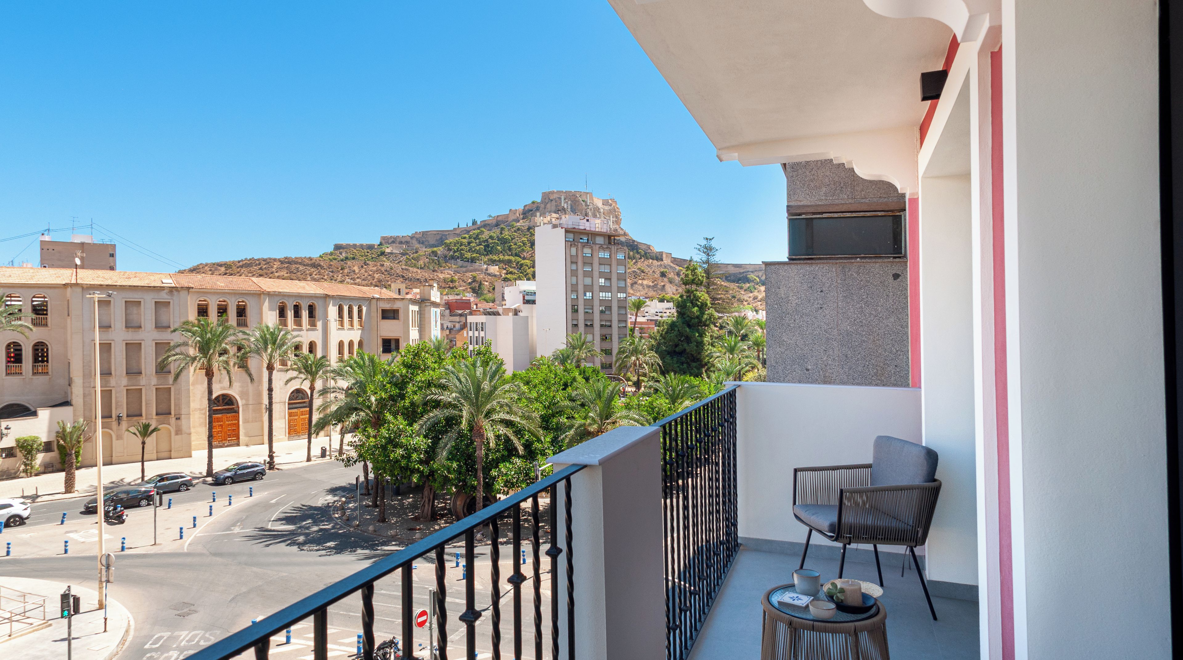 Un balcon avec une chaise et une table, donnant sur une rue bordée de palmiers et d’immeubles, dont un bâtiment imposant situé sur une colline.