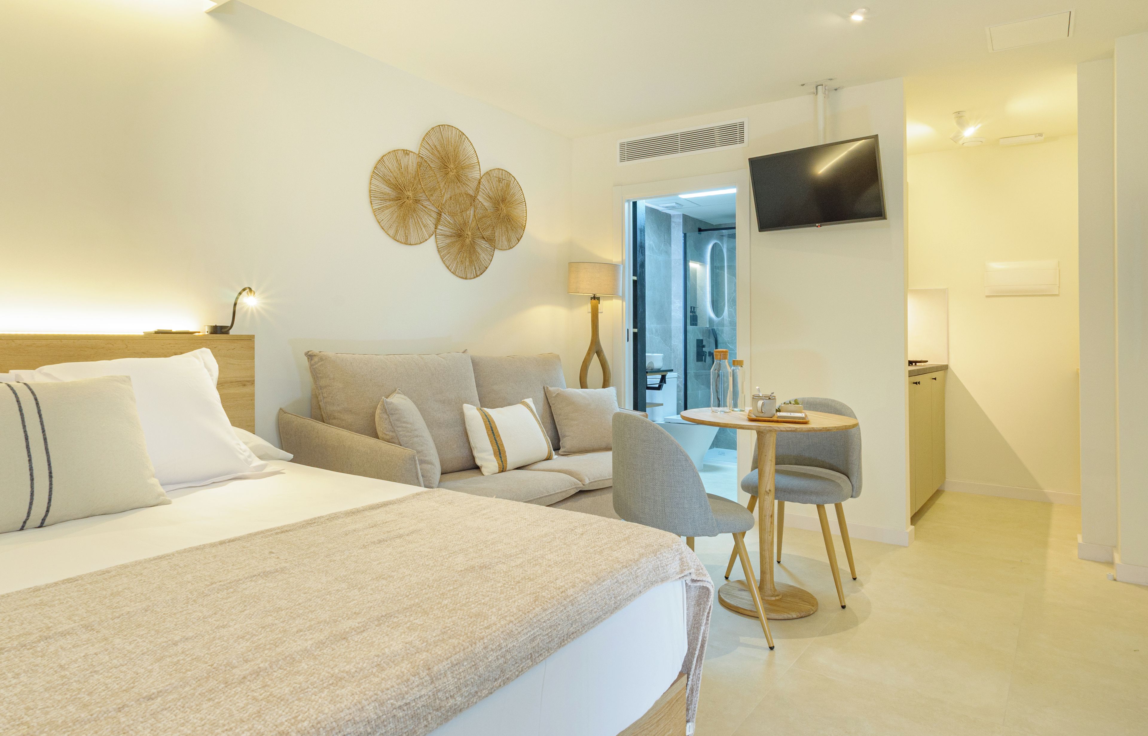 A modern hotel room with a large bed, a sofa, a small table with a chair, a TV mounted on the wall, and a bathroom visible through an open door.