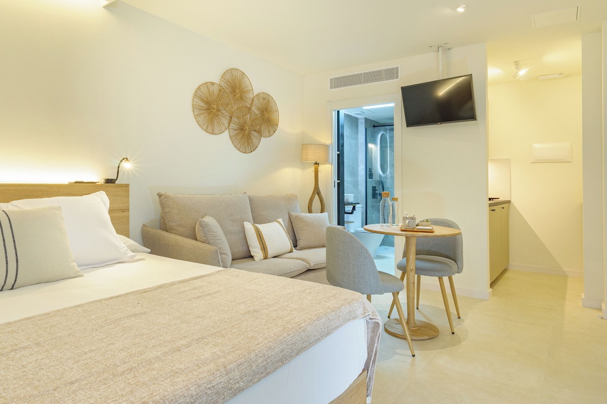 A modern hotel room with a large bed, a sofa, a small table with a chair, a TV mounted on the wall, and a bathroom visible through an open door.