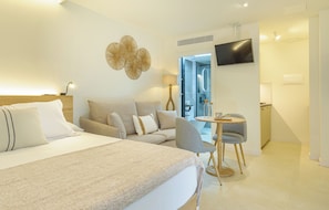 A modern hotel room with a large bed, a sofa, a small table with a chair, a TV mounted on the wall, and a bathroom visible through an open door.