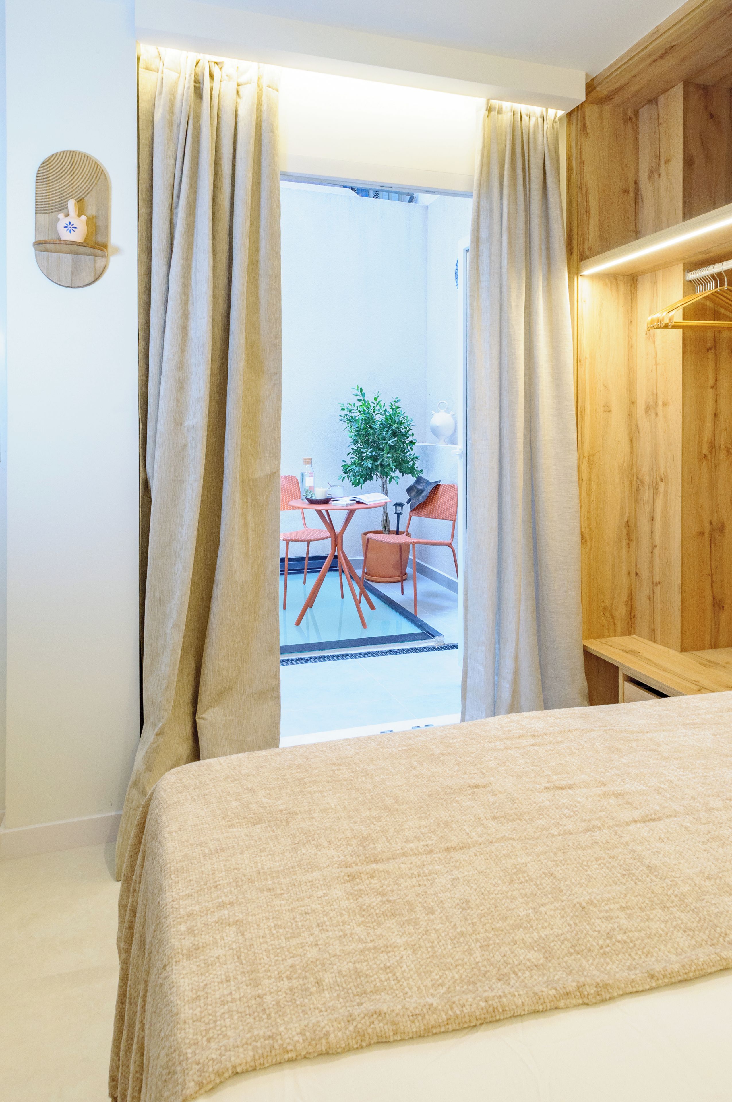 Habitación de hotel con cama, cortinas, un armario de madera y vistas a una zona de estar exterior con mesa, sillas y una planta en maceta.