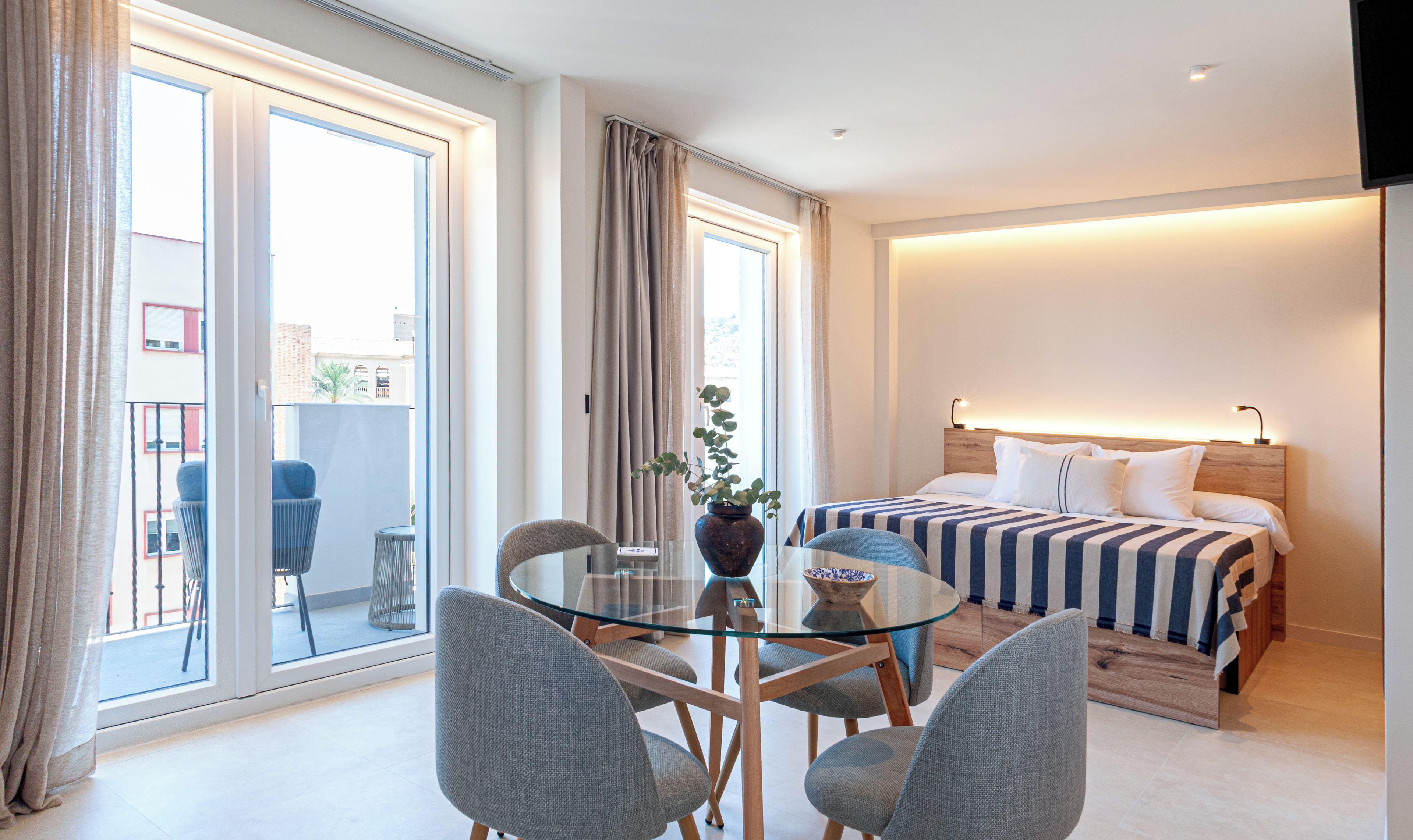 Une chambre d’hôtel avec un lit, une table à manger en verre entourée de chaises, une plante en pot et une vue sur l’extérieur.