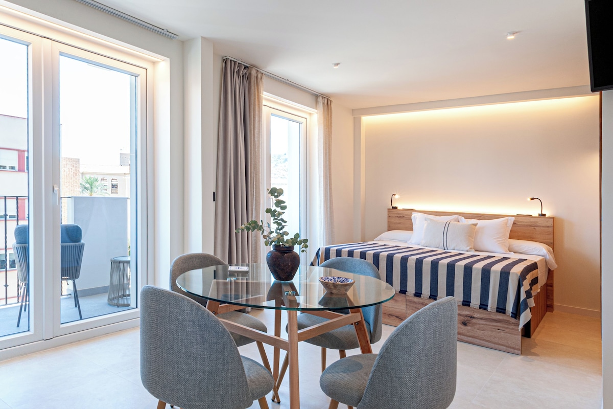 A hotel room with a bed, a glass dining table with chairs, a potted plant, and a view of the outside.
