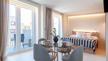 Une chambre d’hôtel avec un lit, une table à manger en verre entourée de chaises, une plante en pot et une vue sur l’extérieur.