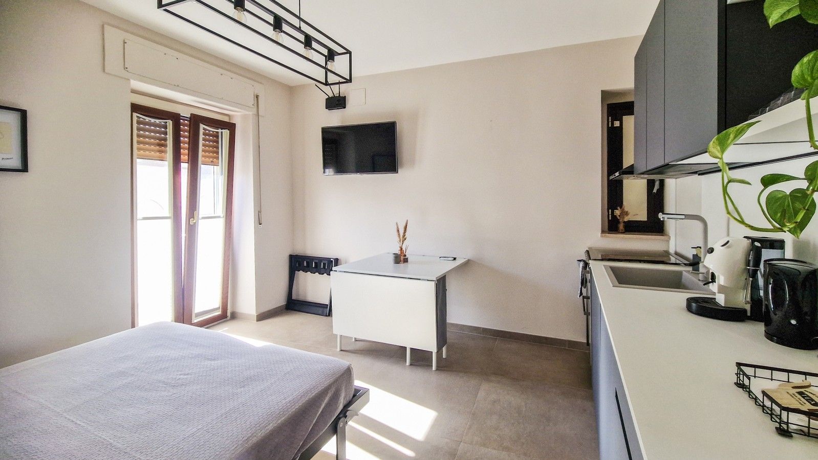 Ein kleines Hotelzimmer mit einem Bett, einem Schreibtisch, einem Fernseher, einer Küchenzeile und einem Fenster mit Jalousien.