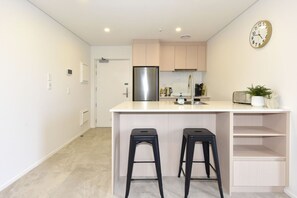 Une cuisine moderne avec un réfrigérateur en acier inoxydable, un plan de travail blanc, deux tabourets noirs et une horloge au mur.