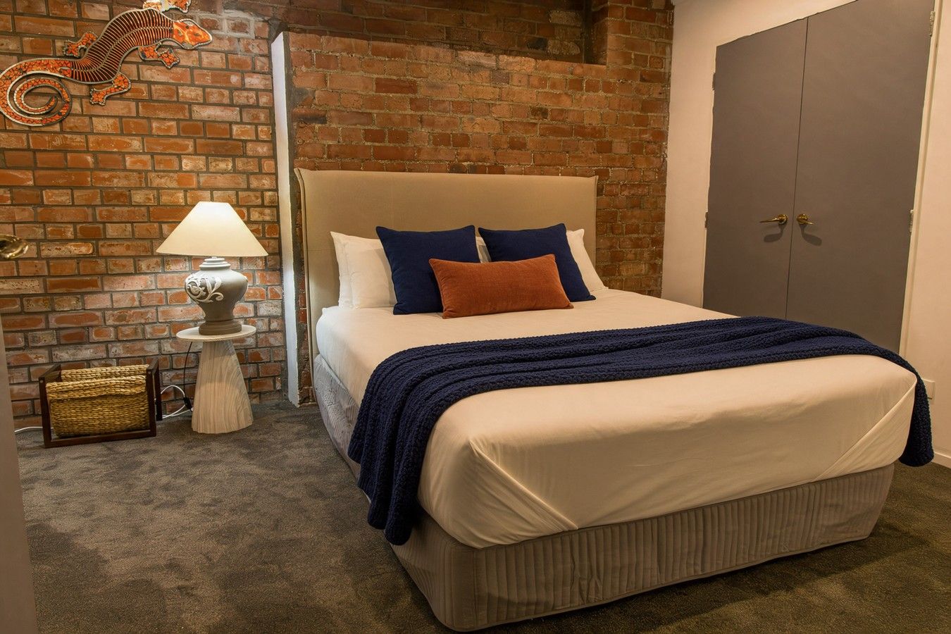 A bedroom with a brick wall, a bed with a headboard, a nightstand with a lamp, and a door.