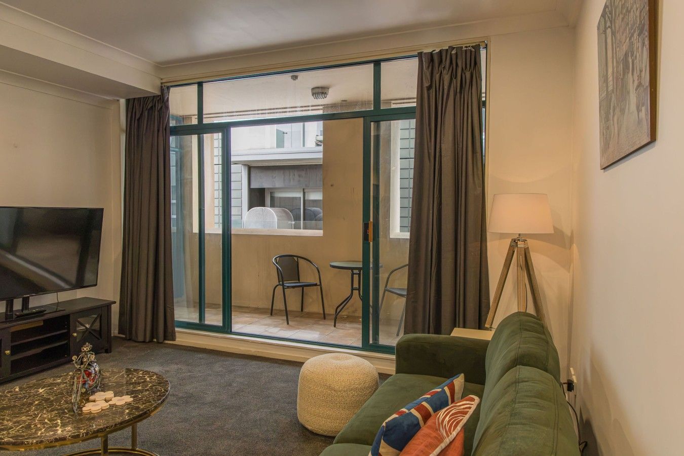 A living room with a television, a sofa, a coffee table, and a view through a glass door to an outdoor area with a table and chairs.