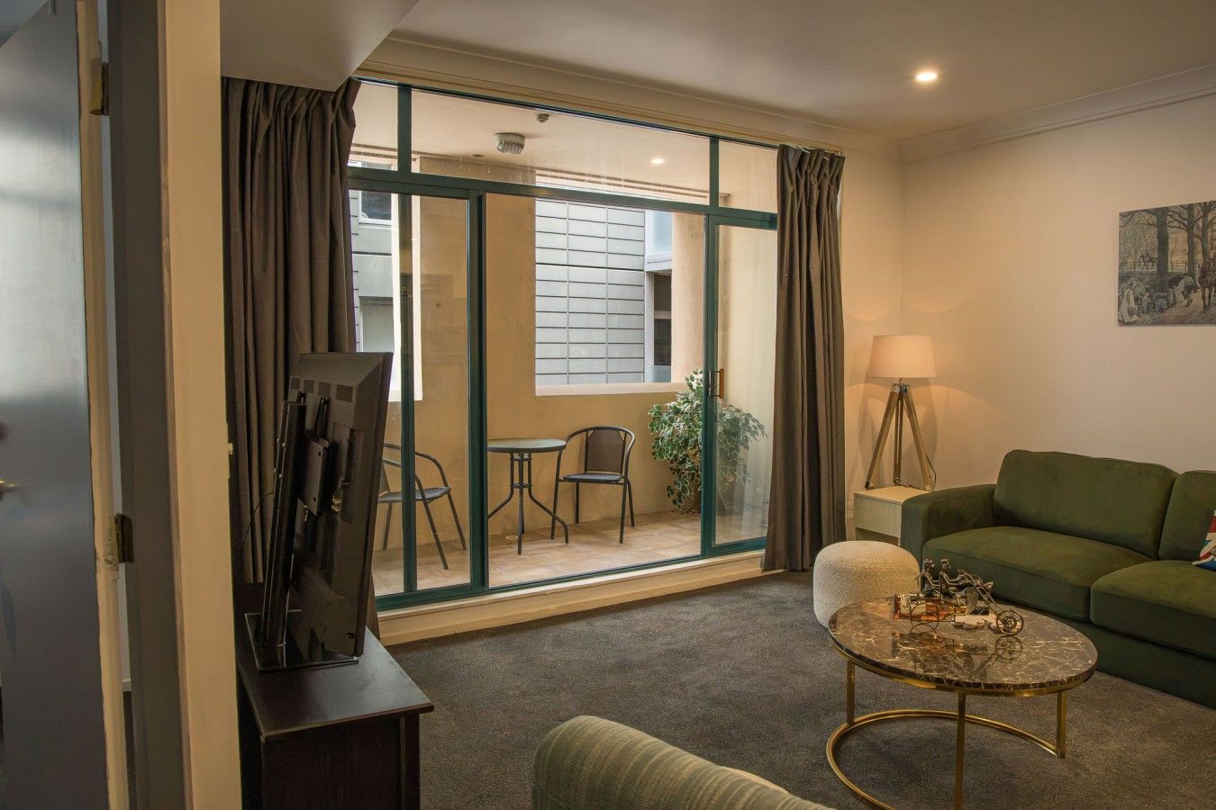 Ein Hotelzimmer mit einem grünen Sofa, einem kleinen runden Couchtisch, einer Lampe und Blick auf den Außenbalkon mit Stühlen und einer Pflanze.