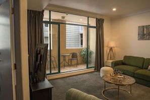 A hotel room with a green sofa, a small round coffee table, a lamp, and a view of the outside balcony with chairs and a plant. - Spacious 3-Bedroom Stay in Central Wellington (Wellington)