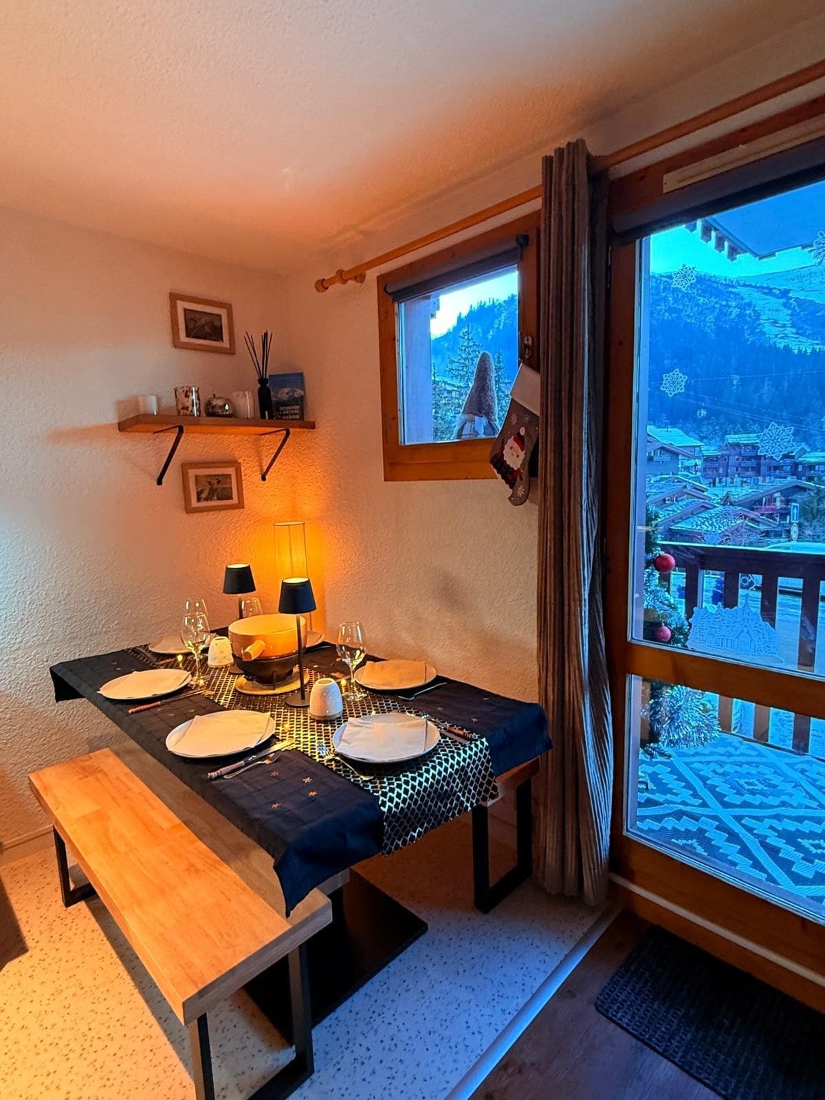 A dining area with a table set for two, a window with a view of a snowy landscape, and a shelf with decorative items.