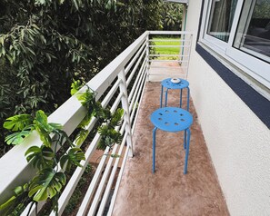Un balcón con barandilla blanca, dos mesas azules y plantas verdes.