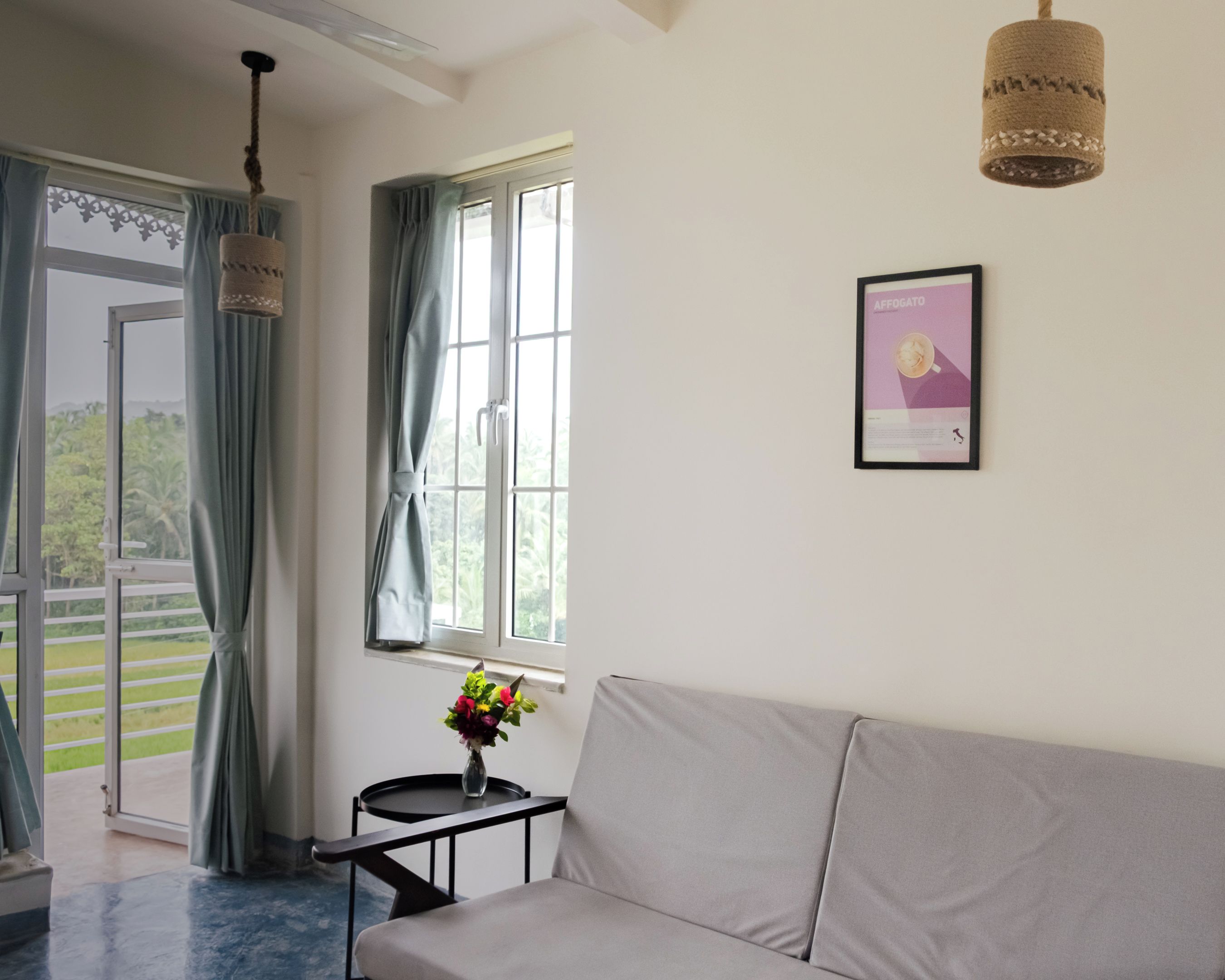 Una habitación con sofá, una pequeña mesa con un florero, un cuadro enmarcado, una ventana con cortinas y vistas a zonas verdes.