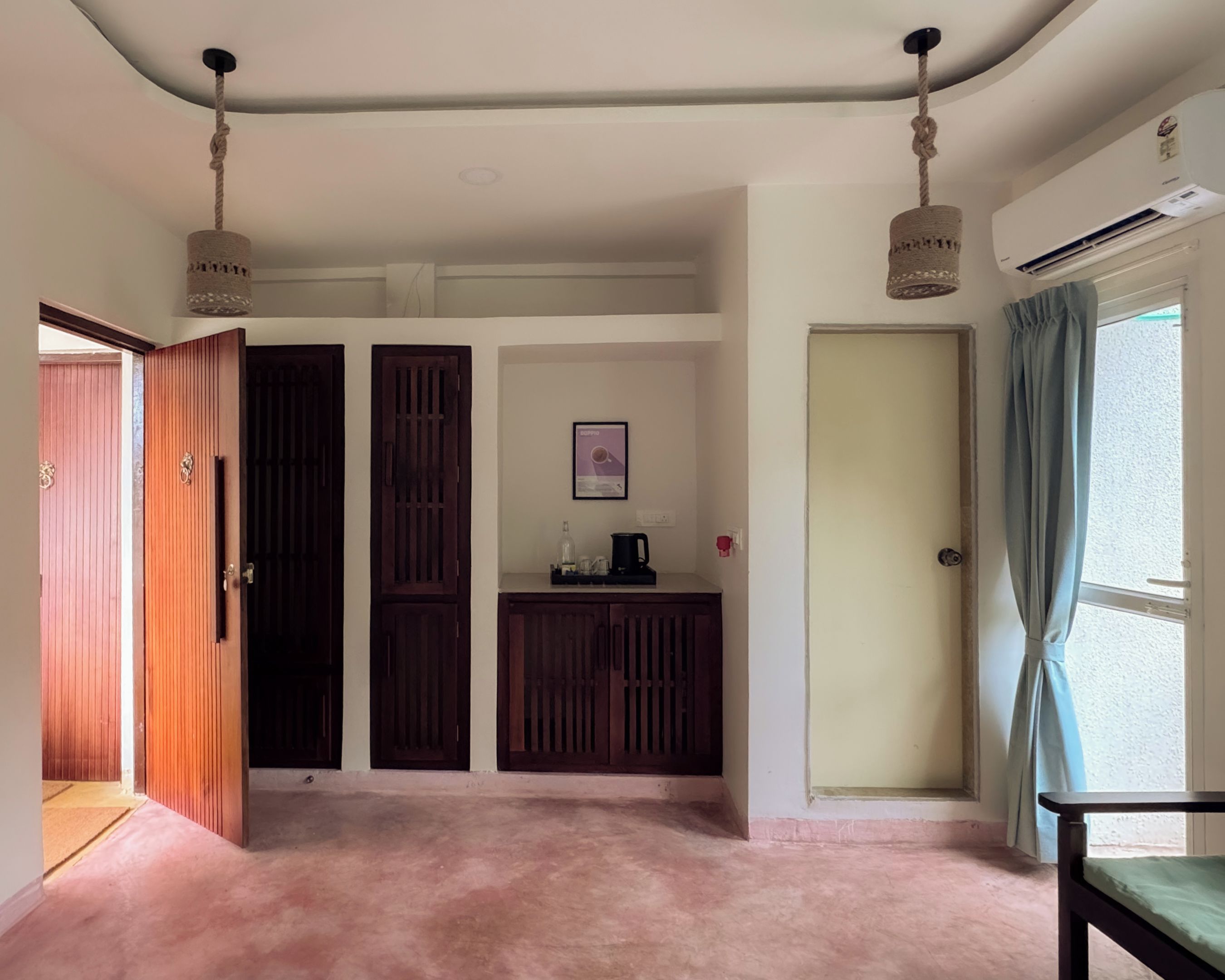 A room with a pink carpet, wooden doors, a small kitchenette, and a window with curtains.