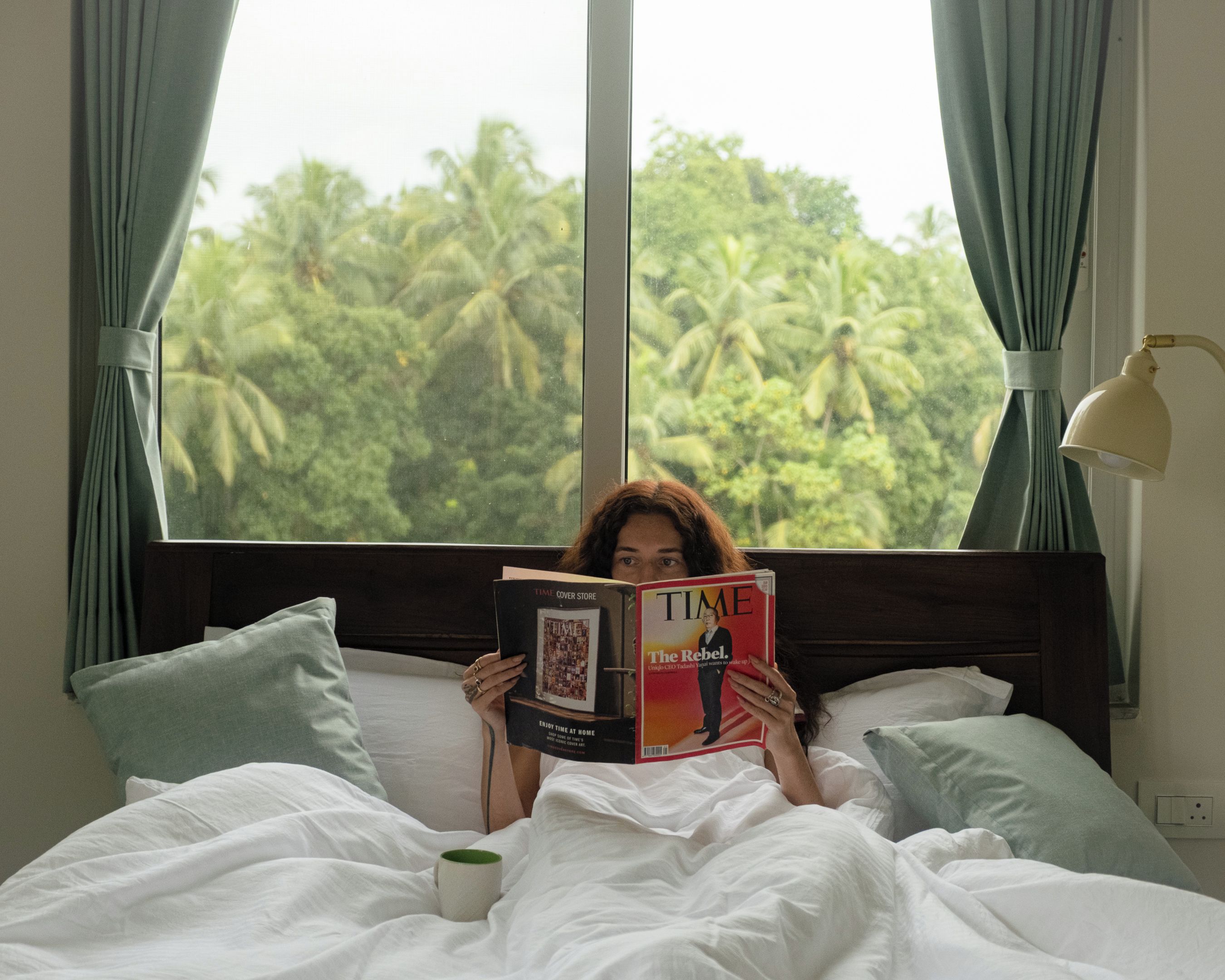 A person lying in bed reading a TIME magazine, with a view of palm trees through a window.