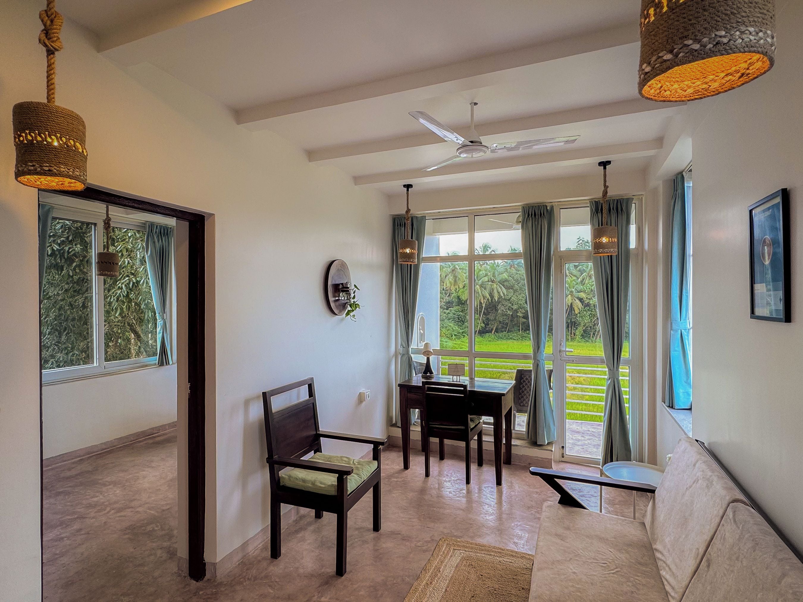 A spacious living area with a ceiling fan, a dining table, a sofa, and large windows offering a view of a lush garden.
