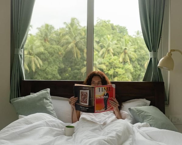 A person reading a book on a bed with a view of palm trees through a window.