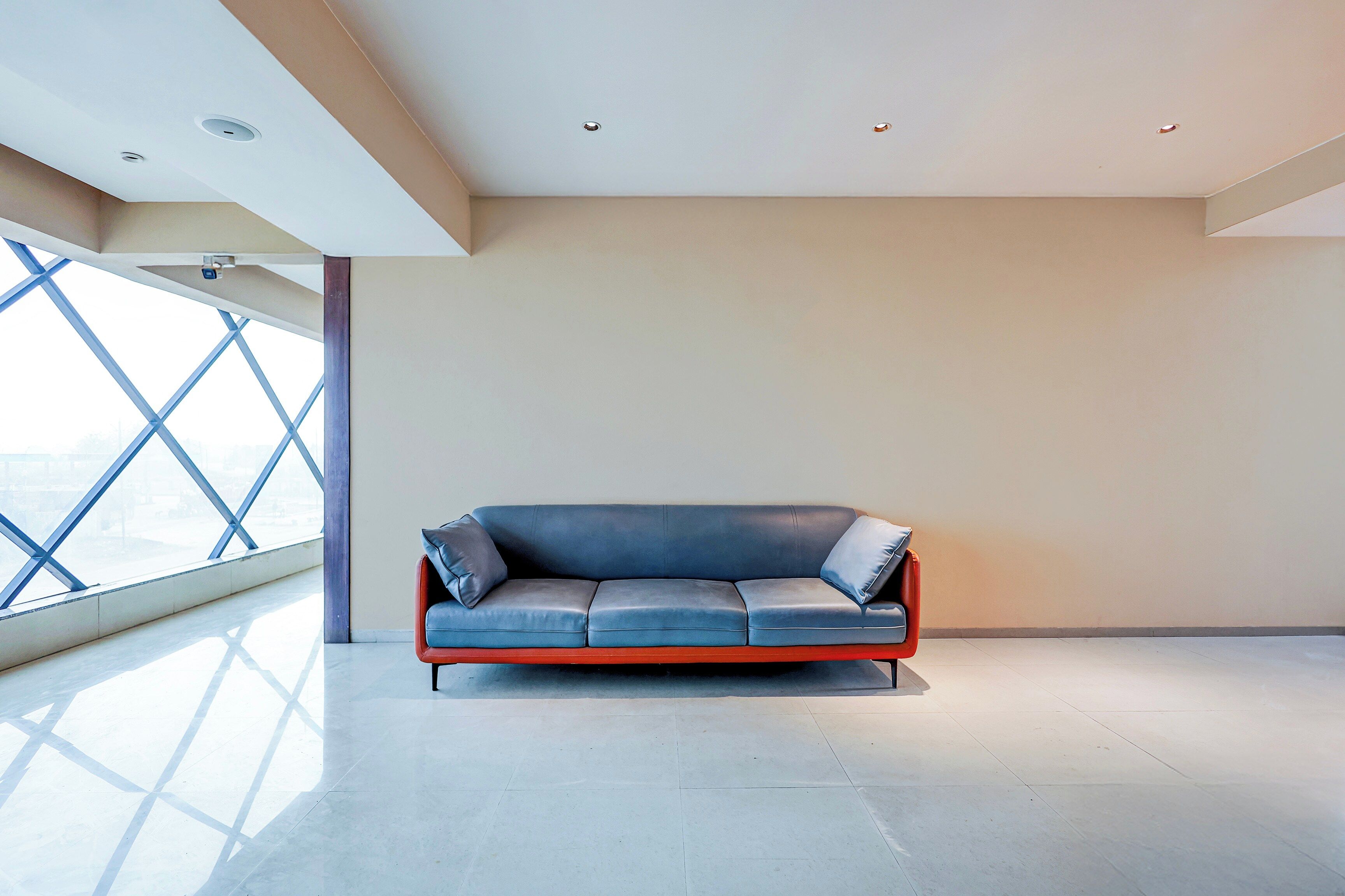 Ein modernes Interieur mit einem einzelnen blauen Ledersofa vor einer schlichten Wand, großen Fenstern und einem geometrischen Deckenmuster.