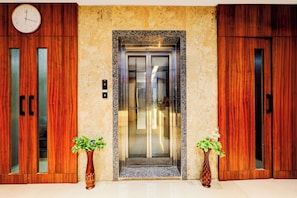 A hotel entrance with a marble wall, a clock, an elevator, and wooden doors.