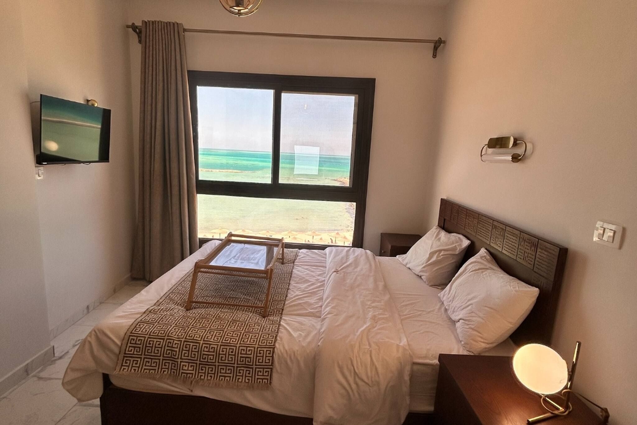 A hotel room with a bed, a bedside table, a lamp, a TV, and a window with a beach view.