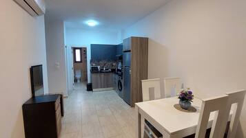 A narrow hallway leading to a kitchen with a microwave, refrigerator, and washing machine. There is a dining area with a table and chairs, and a small window with a view.