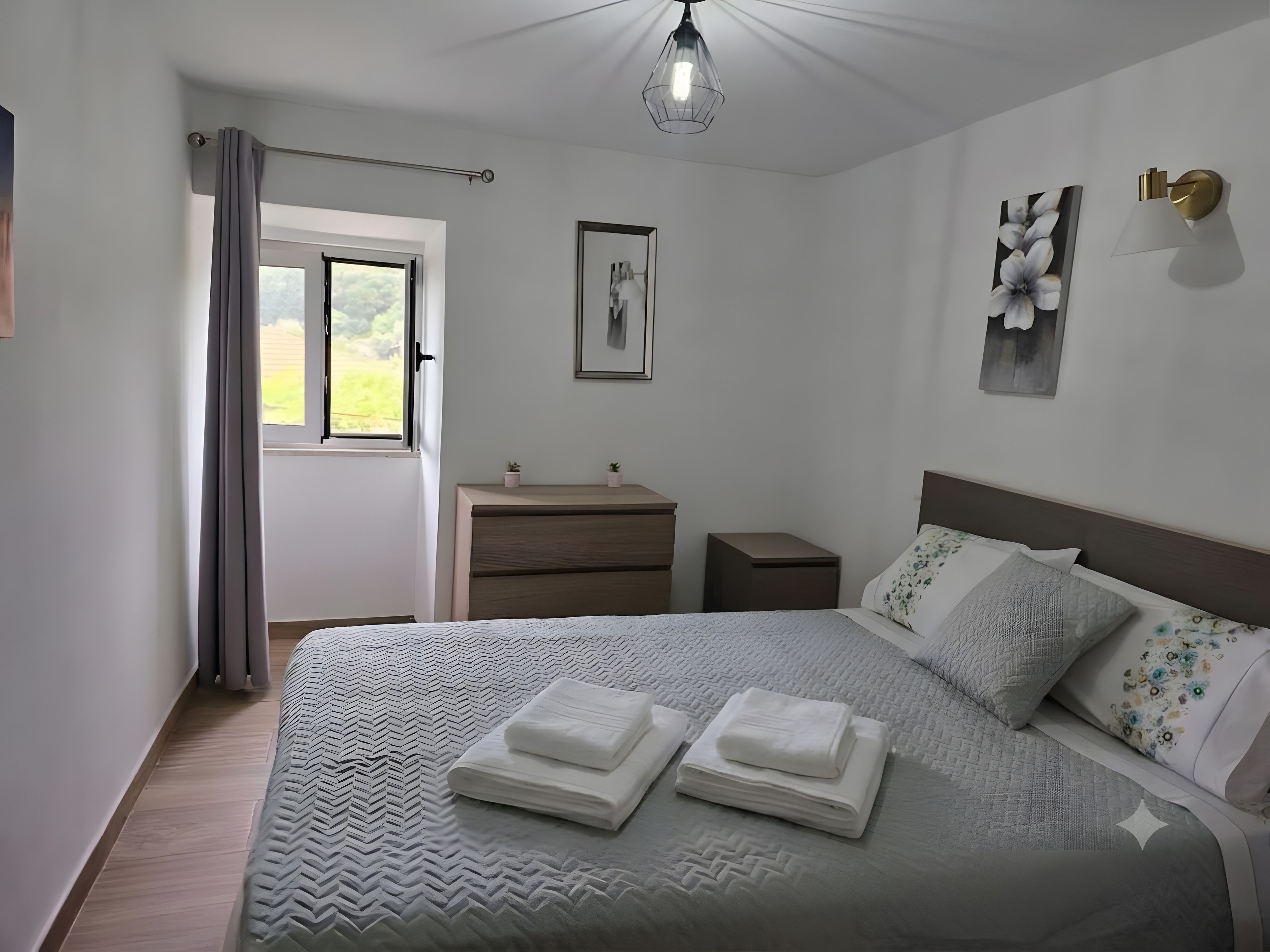 Een net opgemaakt bed met opgevouwen handdoeken, een nachtkastje, een wandlamp en een raam met gordijnen.