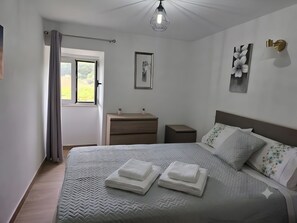 En snyggt bäddad säng med prydligt vikta handdukar, ett sängbord, en liten avlastningsyta bredvid sängen, en vägglampa och ett fönster med gardiner.
