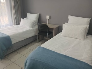 A hotel room with two single beds, a desk, a chair, and a window with curtains. - Spacious 5-bedroom House in Pietermaritzburg With Shared Pool (Pietermaritzburg)