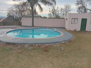 A swimming pool with a curved edge, surrounded by a concrete deck and a grassy area. - Spacious 5-bedroom House in Pietermaritzburg With Shared Pool (Pietermaritzburg)
