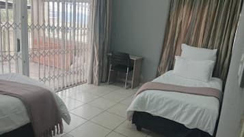 Habitación de hotel con dos camas, un escritorio pequeño y vistas a través de una ventana con barrotes.