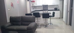 A modern kitchen with white cabinets, a black countertop, and a microwave. There are two black bar stools and a grey sofa in the living area. - Spacious 5-bedroom House in Pietermaritzburg With Shared Pool (Pietermaritzburg)