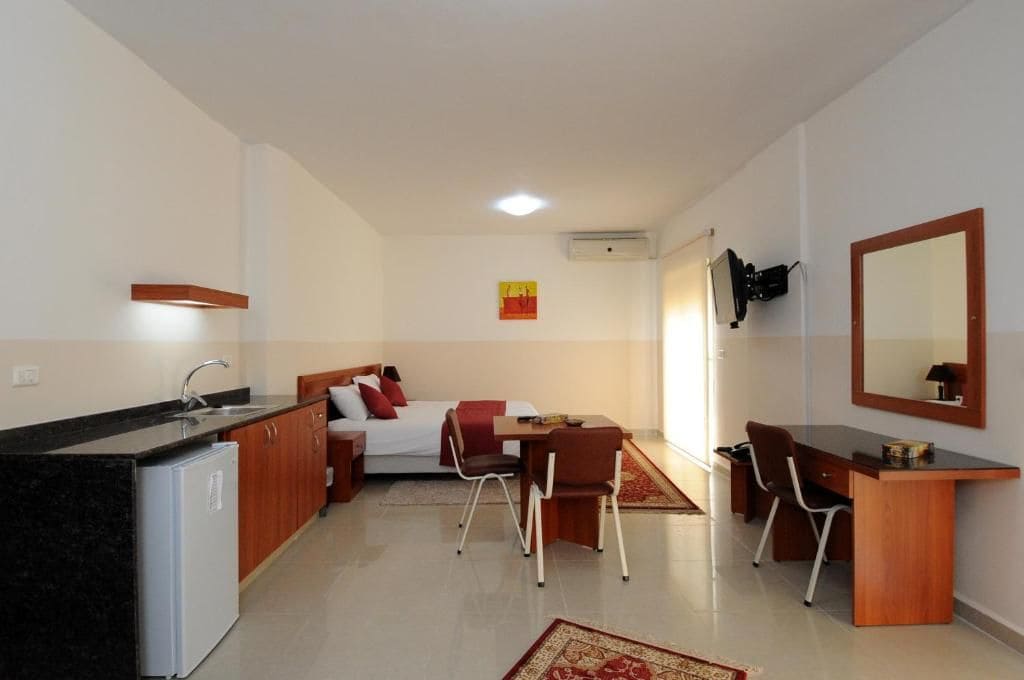 A compact hotel room with a kitchenette, a bed with red pillows, a dining table with chairs, and a large mirror on the wall.