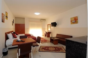 A hotel room with a bed, a desk, a chair, a sofa, a TV, and a kitchen area.