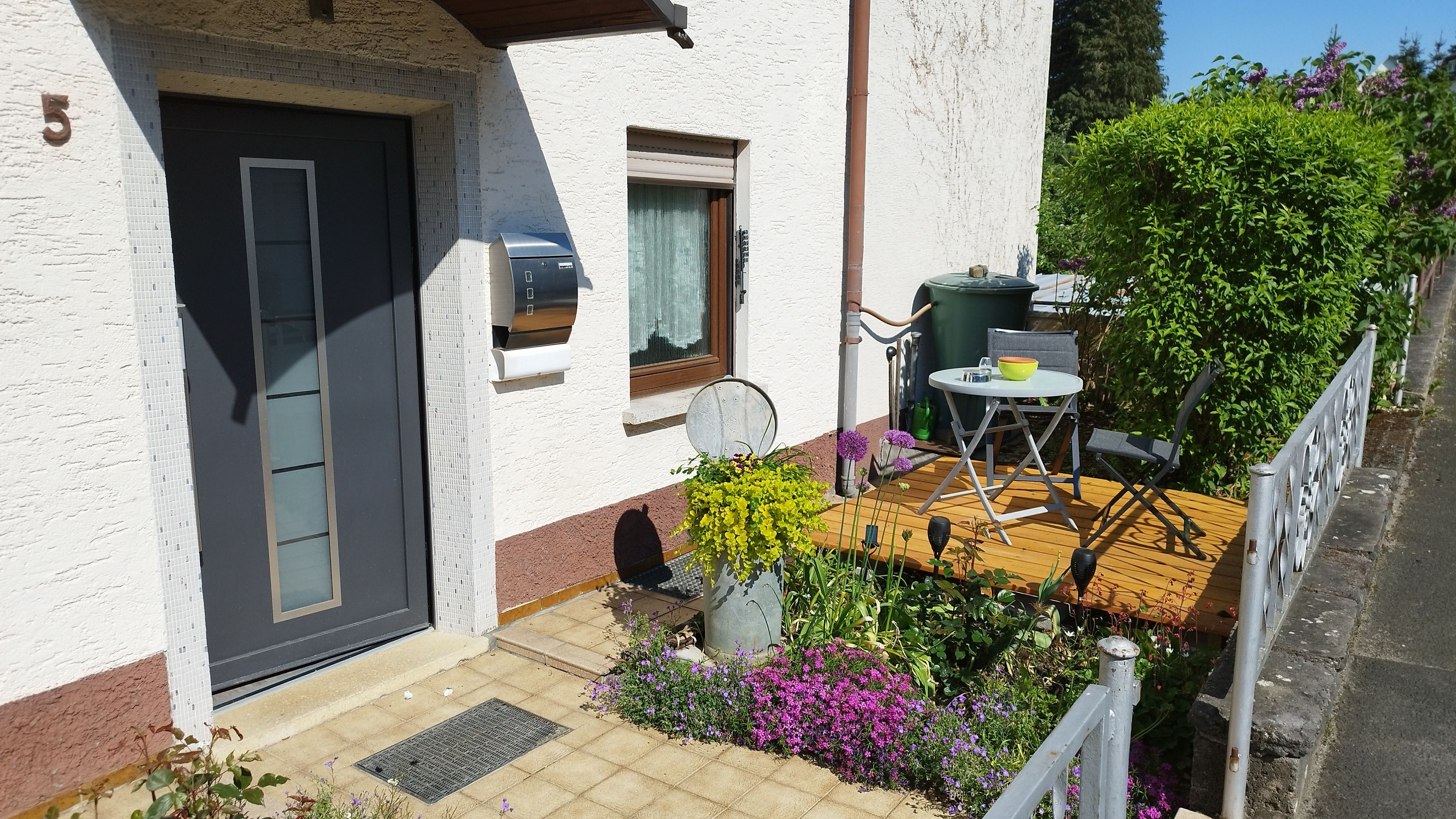 A house with a dark door, a mailbox, a small window with a curtain, a garden with flowers, and a table with chairs outside.