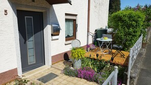 Une maison avec une porte sombre, une boîte aux lettres, une petite fenêtre avec un rideau, un jardin fleuri et une table avec des chaises à l’extérieur.