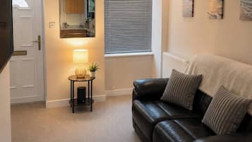 A living room with a black leather sofa, a small side table with a lamp, a window with blinds, and a door leading to another room.