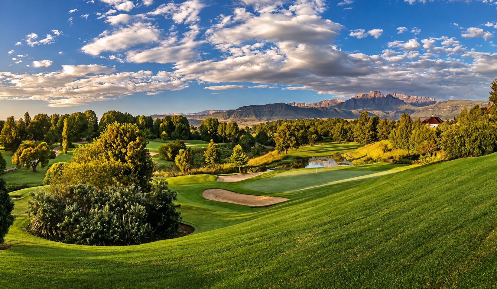 Un campo de golf con un estanque, árboles y montañas al fondo.