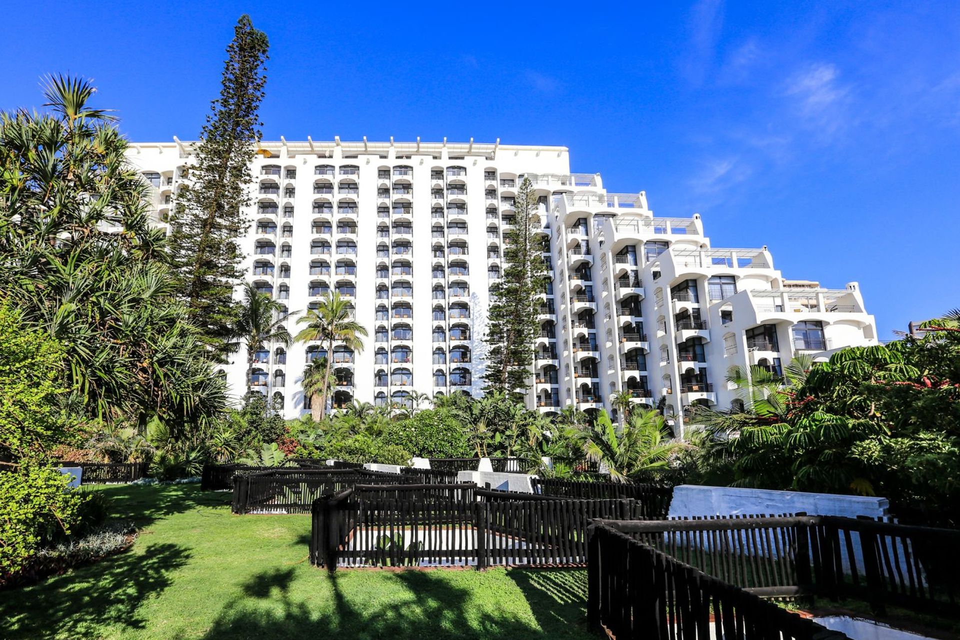 A multi-story hotel with a white facade, balconies, and a well-maintained garden area.