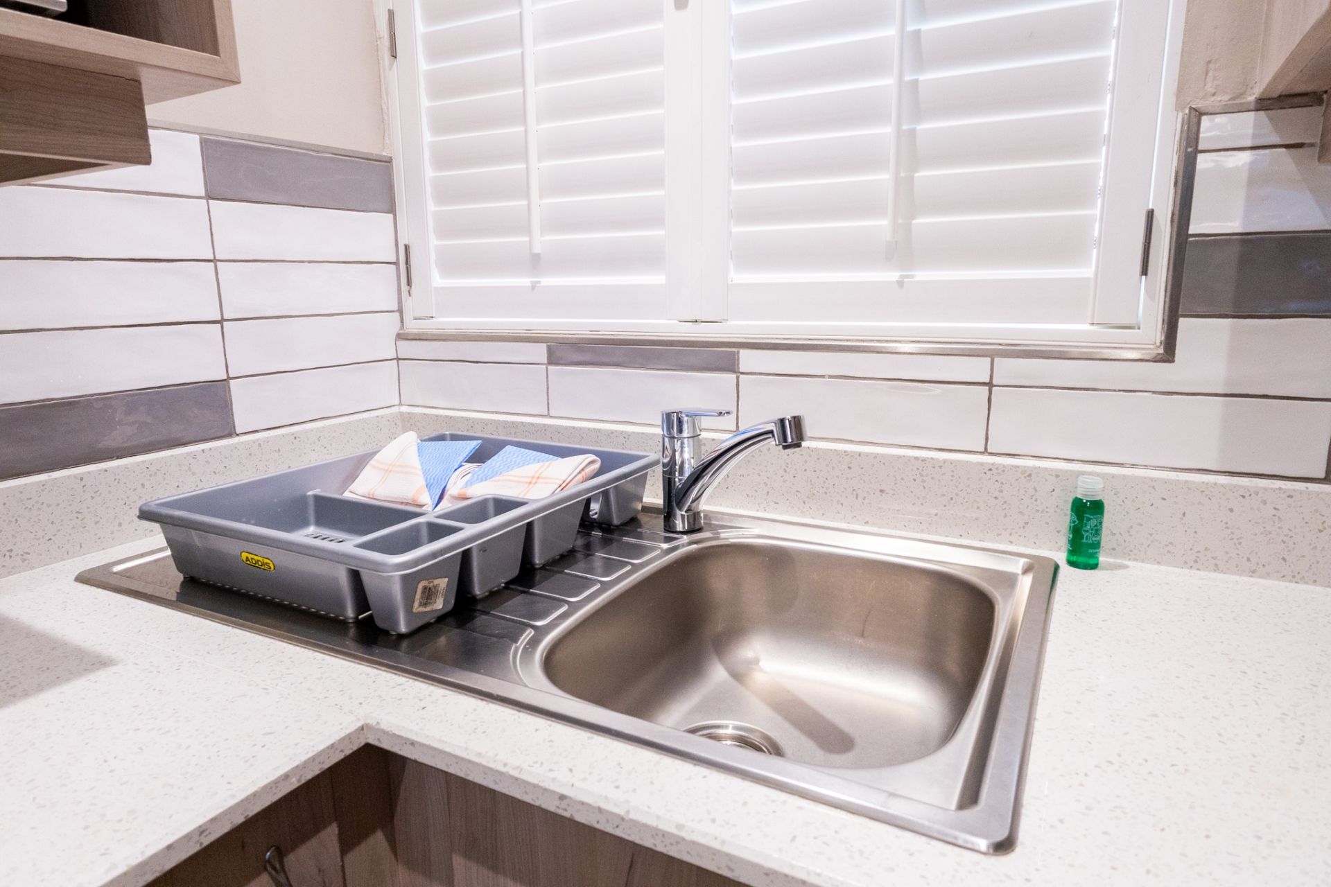 A kitchen with a stainless steel sink, a window with blinds, and a countertop with a dish rack.