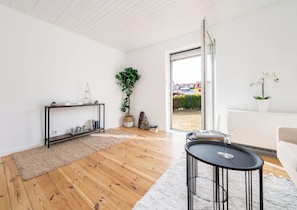 Ein modernes Wohnzimmer mit Holzboden, einem schwarzen Konsolentisch, einem weißen Sofa und einem großen Fenster mit Blick auf Gebäude und Grünflächen.