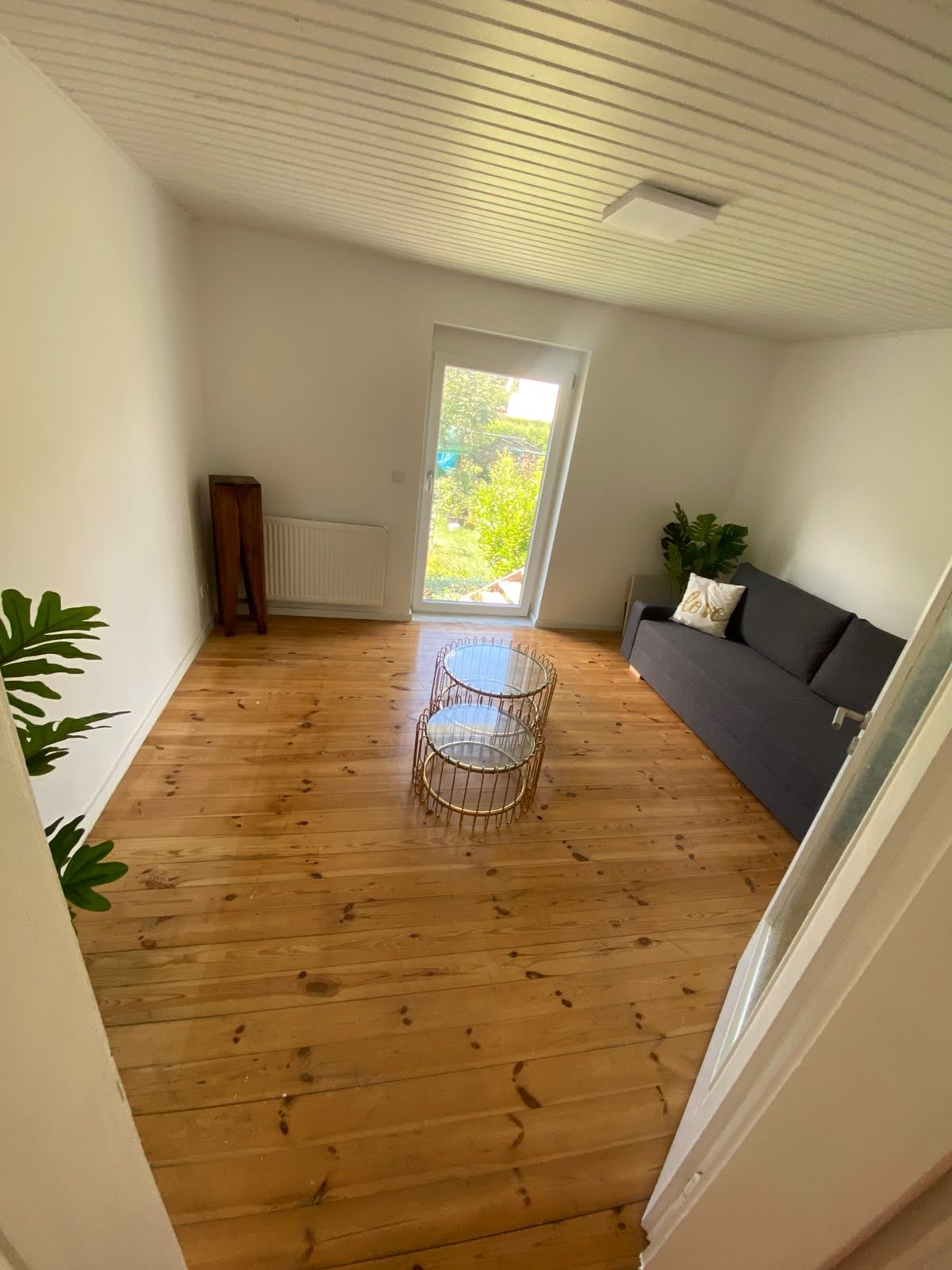 Ein Zimmer mit Holzboden, einem grauen Sofa, einem kleinen runden Tisch und einer Tür zu einem Balkon mit Grünpflanzen.