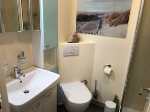 A bathroom with a white sink, a toilet, and a shower. There is a painting on the wall above the toilet. - 10 in Goting Huus (Wyk auf Foehr)