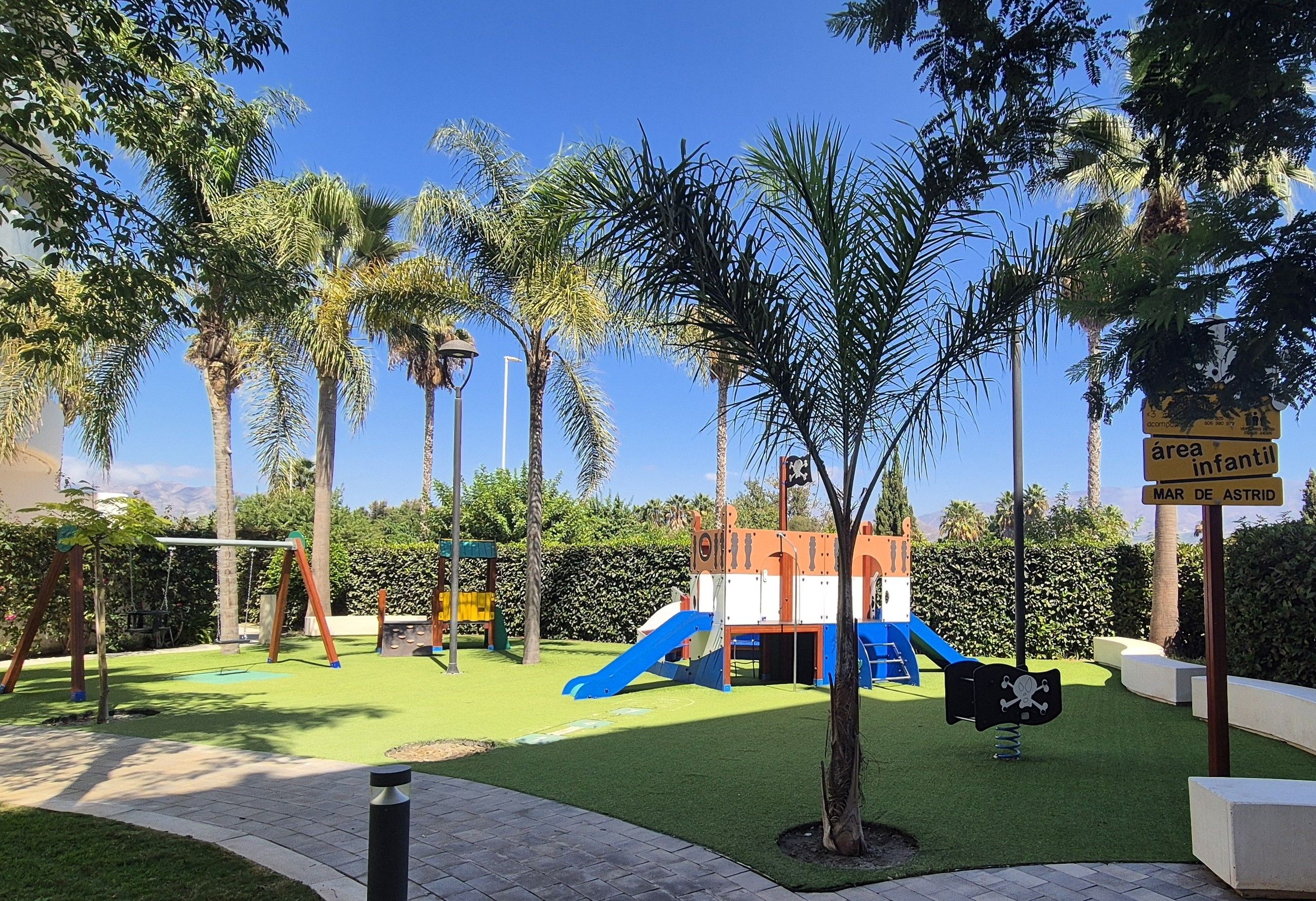 Un parque infantil con toboganes y estructuras de escalada, rodeado de palmeras y un cielo despejado.